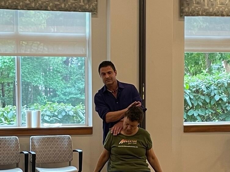 A man stands behind a seated person and applies pressure to their shoulder and neck area in what appears to be a therapeutic 