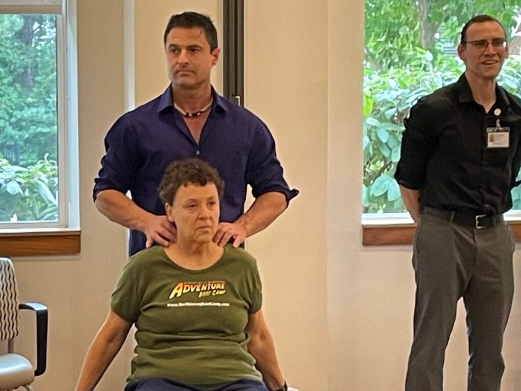 A man stands behind a seated woman, placing his hands on her shoulders in what appears to be a demonstration or therapeutic t