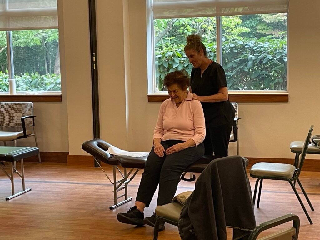A caregiver or therapist provides a shoulder massage to a seated elderly woman in a treatment room